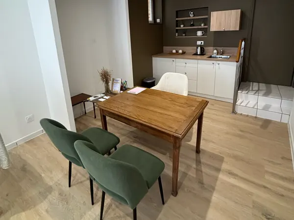 Salle avec table en bois, chaises vertes et coin kitchenette